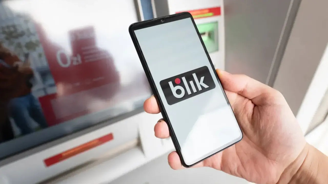 Dłoń trzyma smartfon z logo Blik. Obok bankomat, być może próba płatności Blikiem na zły numer telefonu.