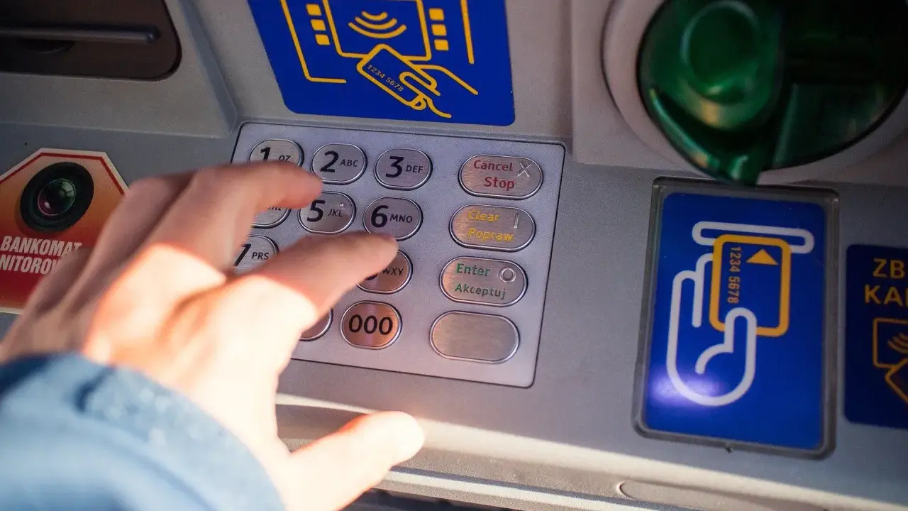 Dłoń wpisuje PIN na bankomacie. Co zrobić, gdy bankomat wciągnął kartę i jak załatwić zwrot?