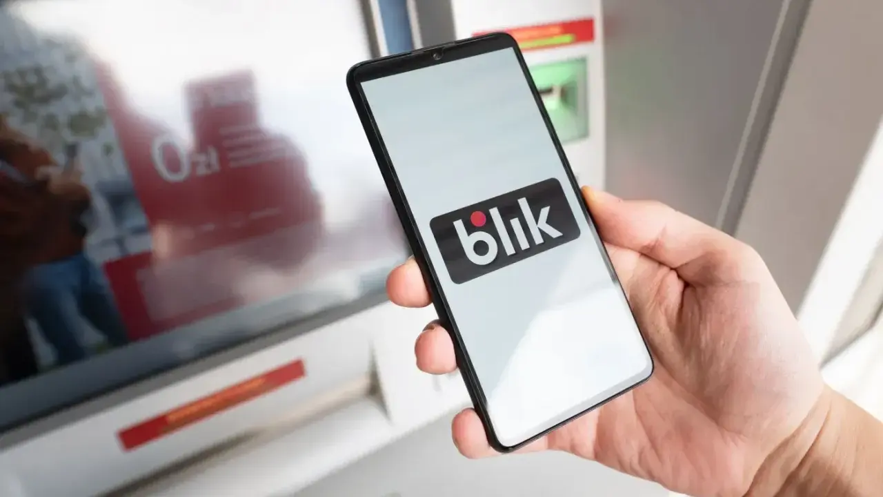 Dłoń trzyma smartfon z logo Blik przed bankomatem. Sprawdź, ile można wypłacić z bankomatu tą metodą.
