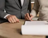 Podpisanie dokumentów dotyczących decyzji o zwrocie podatku z Niemiec. Dwie osoby przy biurku, jedna pisze piórem.