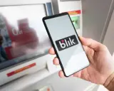 Dłoń trzyma smartfon z logo Blik przed bankomatem. Sprawdź, ile można wypłacić z bankomatu tą metodą.