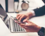 Dłoń trzyma smartfon, obok laptopa. W tle zegar. Szybkie przelewy przychodzące Millennium to wygoda.