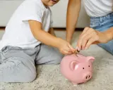 Dziecko i mama wrzucają monety do różowej świnki-skarbonki. Uczą się oszczędzać, by wiedzieć, kto wypłaca rodzinne.