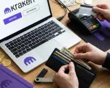Jak wpłacić pieniądze na kartę? Na ekranie laptopa widoczny jest przycisk "Deposit" na platformie Kraken. Obok leżą pieniądze, karty i terminal płatniczy.
