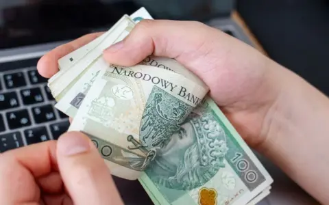 Ręce trzymające stos banknotów 100 zł na tle klawiatury laptopa. Czy w Żabce można wpłacić pieniądze?