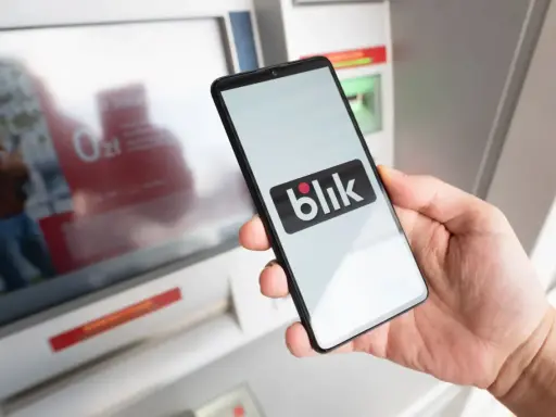 Dłoń trzyma smartfon z logo Blik przed bankomatem. Sprawdź, ile można wypłacić z bankomatu tą metodą.