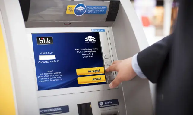 Mężczyzna używa bankomatu do wypłaty pieniędzy telefonem BLIK. Na ekranie widnieje instrukcja "Wprowadź kod BLIK".