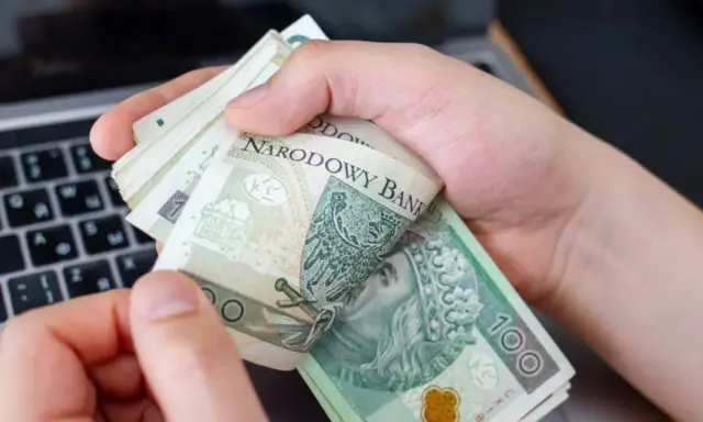 Ręce trzymające stos banknotów 100 zł na tle klawiatury laptopa. Czy w Żabce można wpłacić pieniądze?
