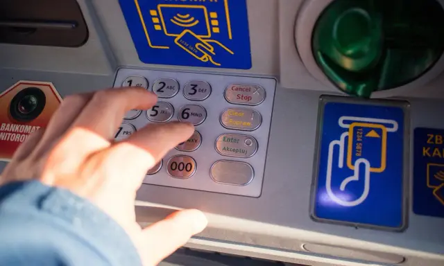 Dłoń wpisuje PIN na bankomacie. Co zrobić, gdy bankomat wciągnął kartę i jak załatwić zwrot?