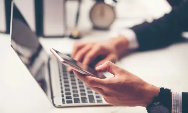 Dłoń trzyma smartfon, obok laptopa. W tle zegar. Szybkie przelewy przychodzące Millennium to wygoda.