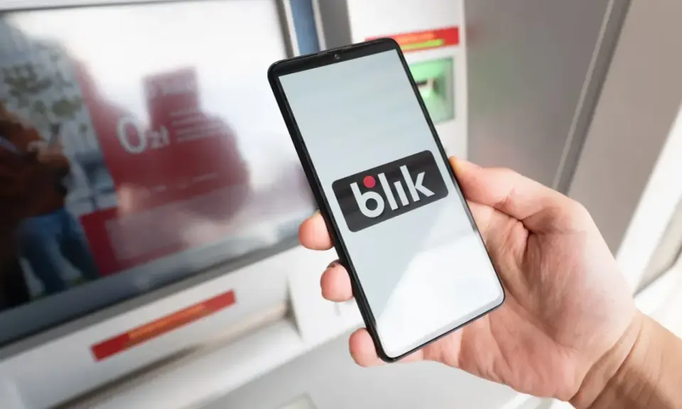 Dłoń trzyma smartfon z logo Blik przed bankomatem. Sprawdź, ile można wypłacić z bankomatu tą metodą.
