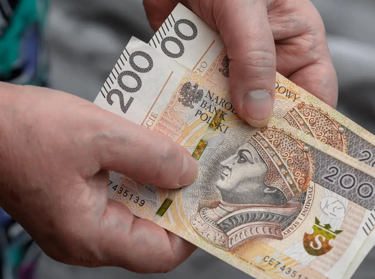 Dłonie trzymają banknoty 200 zł, symbolizujące dodatek do emerytury.