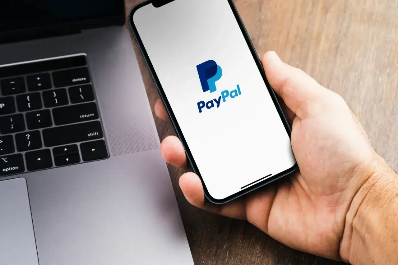 Dłoń trzyma smartfon z logo PayPal, obok laptop. Szybko dowiedz się, jak z PayPal przelać pieniądze na konto.