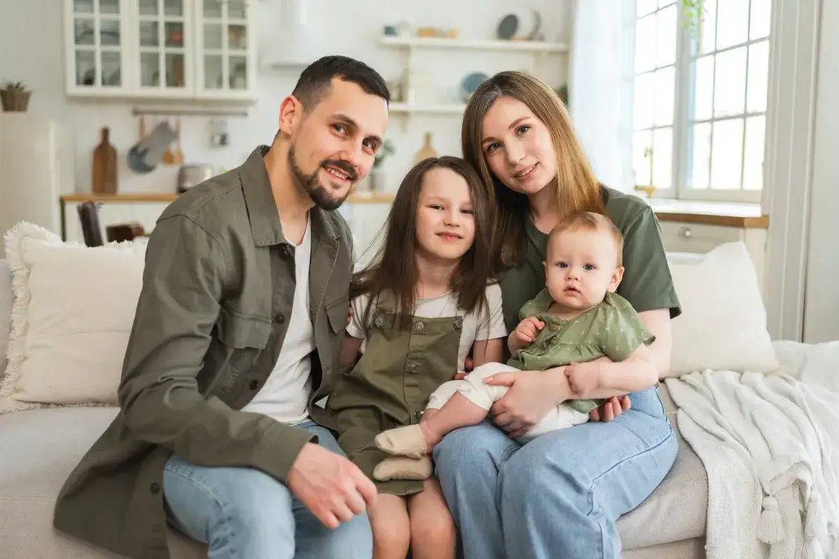 Rodzina na kanapie. Czy 800+ wlicza się do dochodu? Rodzice z dw&oacute;jką dzieci, starszą c&oacute;rką i niemowlęciem, uśmiechają się do aparatu.