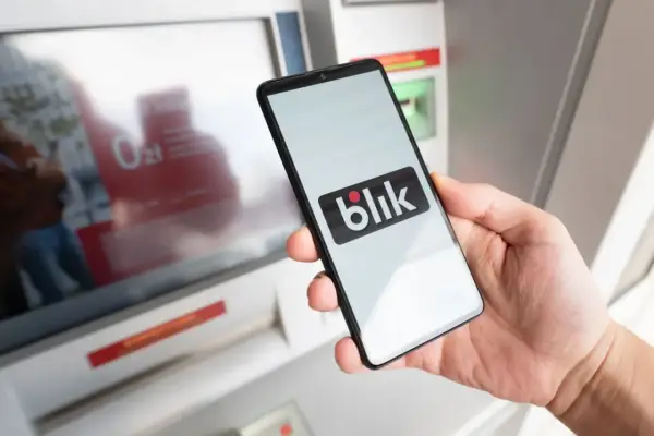 Dłoń trzyma smartfon z logo Blik przed bankomatem. Sprawdź, ile można wypłacić z bankomatu tą metodą.